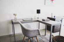 Modern home office with an L-shaped desk, gray upholstered chair, black desk lamp, laptop, cup, and a vase of pink flowers on the desk.