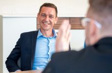 Smiling man in a navy blazer and light blue shirt during a meeting, with a blurred teammate performing a high-five in the foreground.
