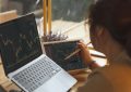 Person uses a tablet and a laptop to monitor stock charts at a wooden desk, holding a pen nearby.