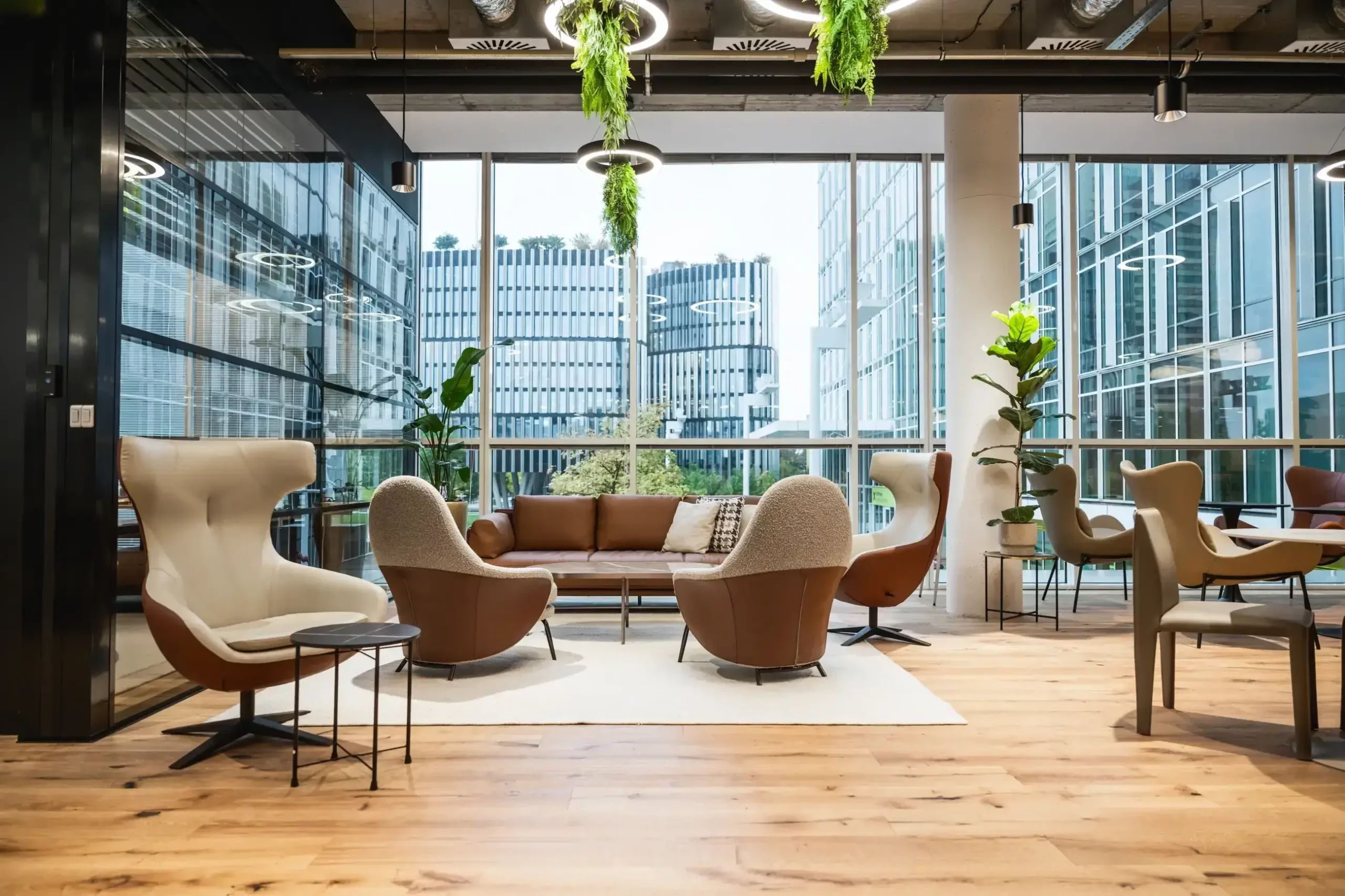 kancelář | Epeníze Modern office lounge with curved beige chairs, a brown sofa, and low tables, near floor-to-ceiling windows and hanging greenery.